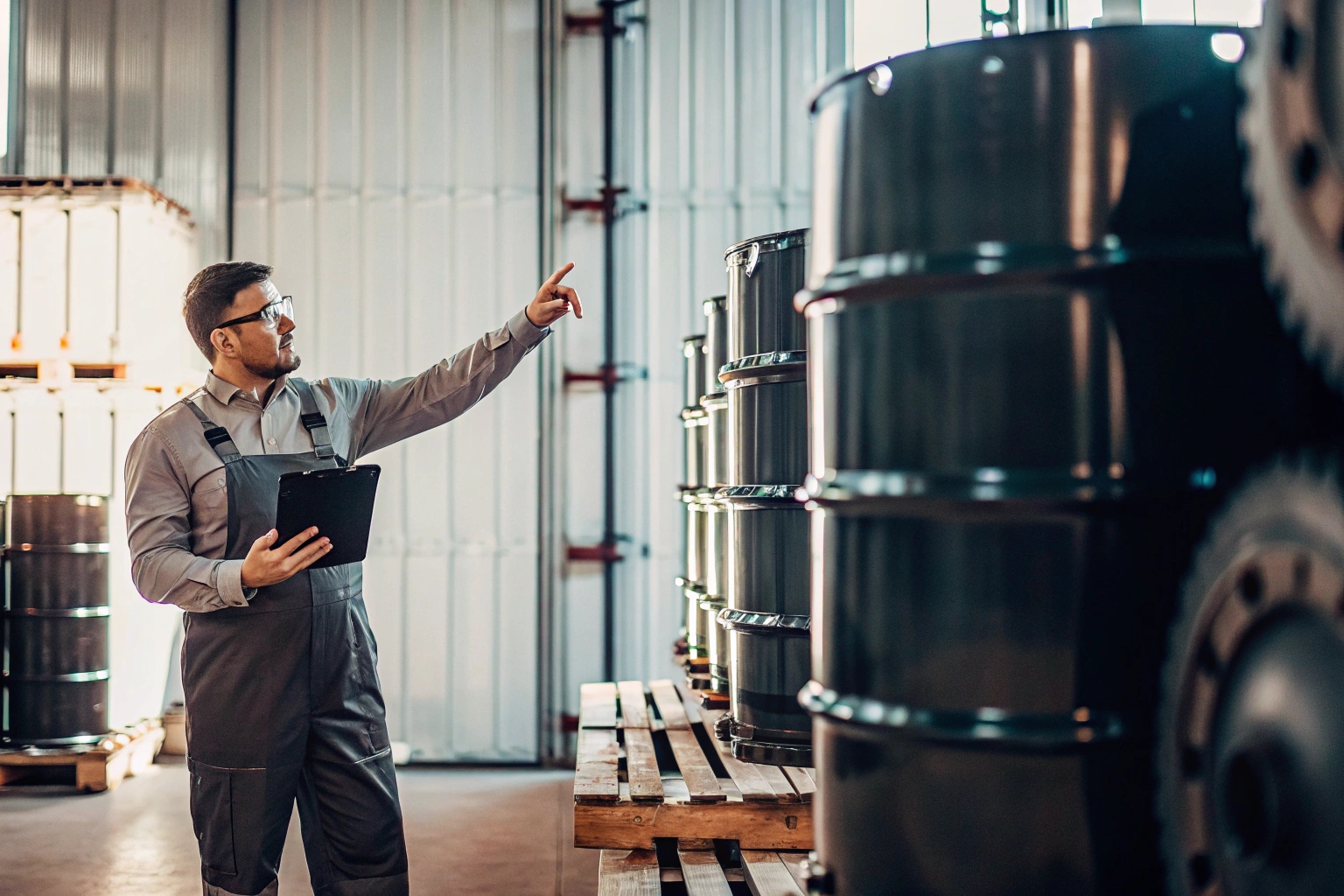 Warehouse worker in overalls holding clipboard and inspecting inventory of black industrial barrels.