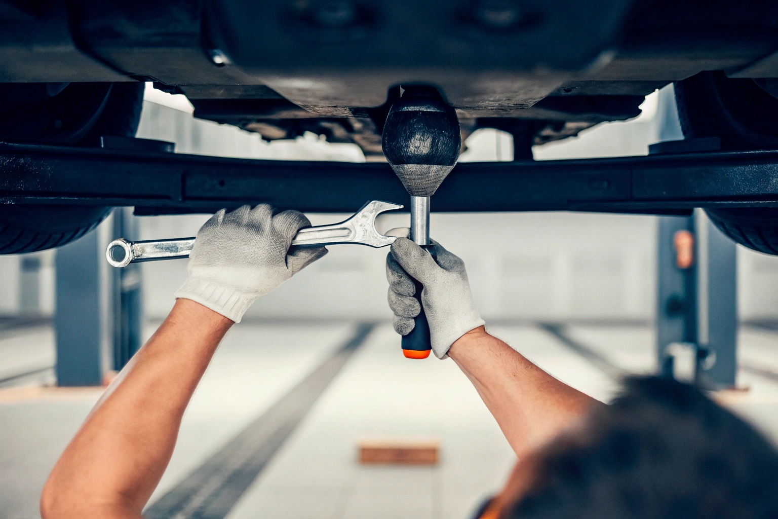 Mechanic in gloves using a wrench to repair car suspension on a lift.